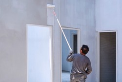 Asian builder using long handle roller brush to painting primer white color on concrete wall inside of house construction site