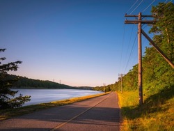 Paved two lane bikeways, electricity poles, white glowing river, blue sky, and winding island forest at sunrise along Cape Cod Canal