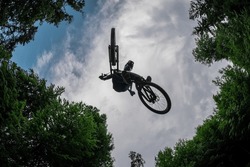 Silhouette of a mountain biker jumping over camera and performing tail whip. Extreme photo of mtb racer jumping surrounded by trees and cloudy sky. Good background and action shot.