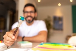 hand showing brush with paint, in blurred background brunette man smiling