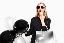 Elegant shocked girl, in black dress with sunglasses, with bags and black balls in her hands isolated on white background . Black Friday, shopping, discounts
