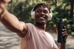 Portrait of smiling african american man wearing headphones taking selfie after jogging holding water bottle outdoors in park