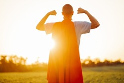 Back view. Successful hero wearing superhero cape, raising arms, showing biceps, showing courage and strength, looking serious and confident, posing against sunset background outdoors