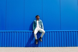 Handsome african male fashion model in sunglasses smiling and posing while standing near blue wall outdoors. Stylish curly african man smiling over isolated blue background