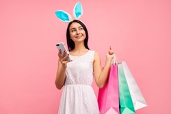 Happy woman in rabbit ears with shopping bags in her hands, typing a message on the phone, traditional holiday, in the studio on a pink background, easter mood