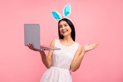 Happy woman in bunny ears holding laptop, video call, shopping online in studio on pink background, easter, christian holiday in april
