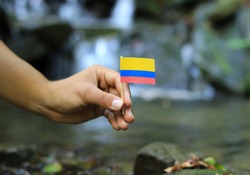 Young man holds national flag of Colombia on wooden stick. Teenager gives respect to nation state of Colombia. Stream and beauty of nature in background. Concept of humanity and prosperity.
