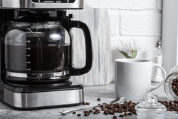 A black drip electric coffee machine with a glass teapot brews a morning drink. Household appliances, a white cup and a jar of beans on the kitchen table on the countertop at home