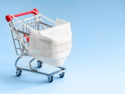 Shopping cart with protective medical mask against coronavirus. Safe and online shopping on quarantine concept. Light blue background, copy space.