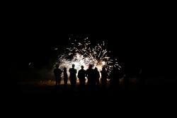 Group of people in silhouette watch fireworks explode on beach in total darkness.