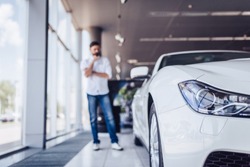 front view of white car in dealership salon with pensive man on the background, blurred shot