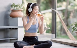 Asian beautiful and sportive woman drinking hot tea and wearing headphone while doing meditation in garden at morning time. Healthy Concept.