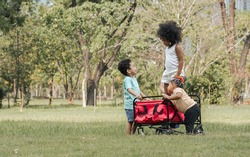 Mixed race little cute children pull and drag a red cart while playing in outdoor green park. Education, Diversity and Teamwork Concept