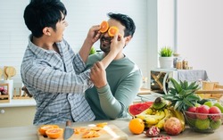 Gay LGBT sweet happy Asian couple wearing pajamas, smiling, taking care, teasing with piece orange on eyes, eating, healthy fruit for breakfast in kitchen at home in morning. Lifestyle, Love Concept