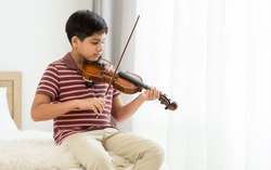 Indian handsome teenage boy, playing, practicing violin musical instrument with happiness in bedroom at cozy home in leisure time, smiling, having copy space. Education, Lifestyle, Creativity Concept