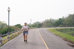 Front view, wide shot with copy space of an active fat young Asian woman with eyeglasses in sportswear running along a curvy road. Sport, weight loss, challenge, self-resolution concept.