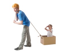 Sad boy with red hair and a blue T-shirt is dragging a cardboard box with little girl sitting in it and showing a hand in the distance on a white background
