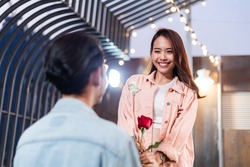 Asian man holding rose standing on one knee to propose his girlfriend on rooftop, focus on happy surprise woman face, romantic dating on Valentine's, anniversary or birthday, Love and marriage concept