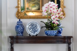 Blue paint porcelain collection with Orchid vase on a dark wooden table with golden mirror on white wall.