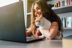 Happy young caucasian woman with her cat using laptop at home. Smart working and smart studying with pet