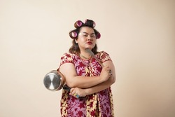 Asian Crazy Housewife. Angry Woman Raising Frying Pan Threatening  Looking At Camera Standing Over creame Studio Background. Furious Wife Shouting Tired Of Cooking Concept