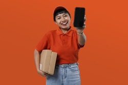 Potrait of young asian courrier woman wearing a red cap and uniform while holding a smartphone and a brown package. Female delivery courier working for a delivery mobile app