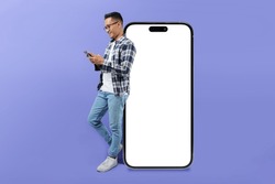 Photo of asian man using mobile phone while standing near huge smartphone with empty screen over light purple studio background. Mockup for website or application, space for advertisement