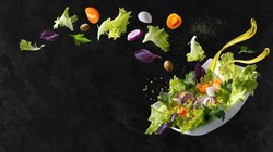 A white plate with salad and floating in the air ingredients: olives, lettuce, onion, tomato, mozzarella cheese, parsley, basil and olive oil. Black background. Copy space.