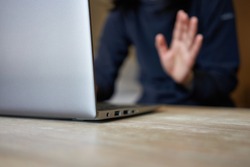 Young woman working from home uses a laptop computer at home to join a video conference meeting and waving hand to her colleagues. Selective focus on the edge of the laptop.