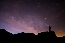 Silhouetted person stands atop boulder with light beam looking up at the milky way and sunrise.