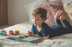 Child reading book. Kids read books. A little boy sits on a bed with your toys in living room watching pictures in story book. Children study.
