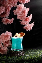 Blue Cocktail decorated with apple form above and dry orange slice. Served on nautre backgorund with moss and  pink blossoms .