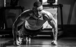 Muscular man doing push-ups on one hand against gym background. Man doing push-ups. Muscular and strong guy exercising. Slim man doing some push ups a the gym. Black and white.