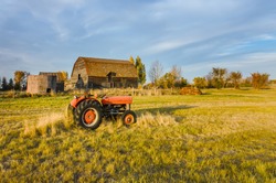 Farm in Saskatchewan, Canada
A red tractor on the grass of a green field, near an old hut and a grain barn with a wooden hedge and a haystack. Trees and blue sky in the background.