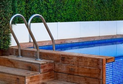 The planking of a swimming pool with stair and turquoise clear water. Open air swimming pool with wooden deck and metal stainless stair and blue surface background. Steps into the pool with handrails