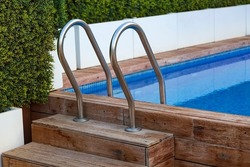 Wooden steps into the pool with metal handrails. The planking of a swimming pool with stair and turquoise clear water. Open air swimming pool with wooden deck, stainless stair and blue surface