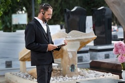 Bearded Jewish man wearing black kippah (skullcap) reading a Hebrew Bible, praying. Prayer in jewish cemetery in Israel. Old jewish cemetery in the forest. A moment from the hasidic jew reading Torah
