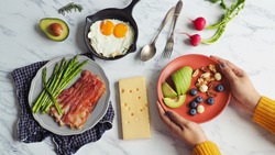 Ketogenic diet meal preparation female hands serving red plate with avocado, blueberry, Brazil nut, macadamia, pecan, walnut on marble table with fried eggs, bacon, roasted asparagus and cheese.