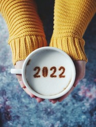 Number 2022 on frothy surface of cappuccino served in white cup holding by female hands over rustic blue background. Holidays food art theme Happy New Year 2022, New year new you. (selective focus)