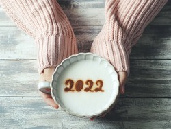 Number 2022 on frothy surface of cappuccino served in pastel pink cup holding by female hands over grey painted wood planks table. Holidays food art theme Happy New Year 2022, New year new you.