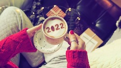 Number 2022 on frothy surface of cappuccino served in white cup holding by woman in red knitted sweater relaxed sitting on the couch. New year new you, Holidays food art theme Happy New Year 2022.