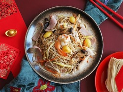 Chinese Lunar New Year lucky food longevity noodle or stir fried noodles served on red background with red envelopes, chopsticks, ancient gold bullion nugget and uncooked dried noodles. (top view)