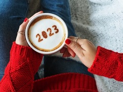 Happy New Year 2023 holidays food art theme, woman in red knitted sweater with jeans holding white coffee cup with number 2023 on frothy surface while relaxed sitting on the couch. (top view)