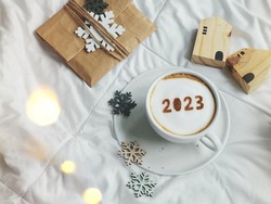 White coffee cup with number 2023 over frothy surface flat lay on the bed with white blanket, wooden house model, paper gift bag, snowflake sign. Home 2023, Happy new year theme (top view, copy space)