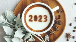 Happy New Year 2023 theme coffee cup with number 2023 over frothy surface served on wooden saucer and white cement background with coffee beans, star anise, silvery gray foliage of dusty miller plant.