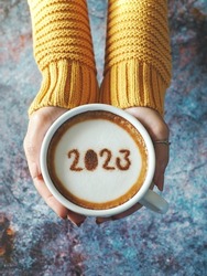 Happy New Year 2023 theme number 2023 on frothy surface of cappuccino served in white coffee mug holding by female hands over rustic blue background. Holidays food art, new year new you. (top view)