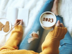 New Year's goal setting, number 2023 on frothy surface of cappuccino in white coffee cup holding by woman in yellow knitted sweater with jeans sitting on bed while writing down her resolutions.