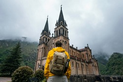 
Young man backpacker with a yellow jacket and mask next to the Sanctuary of Covadonga, Santa Maria la Real, on a cloudy day.