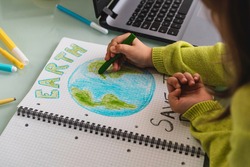 Wide angle view of child girl draws planet earth with wax colors on school notebook for Earth day - Little activist girl writes the message Save the Planet - Protection of environment, global warming
