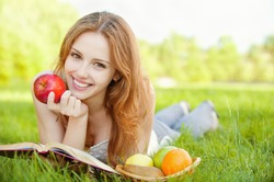 A beautiful young girl with an apple lying on the grass and reading a book beside a basket of fruit is, on a background of green nature